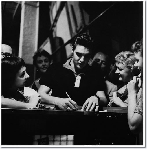 Elvis signs some autographs with British comedienne Joyce Grenfell seen in the background.