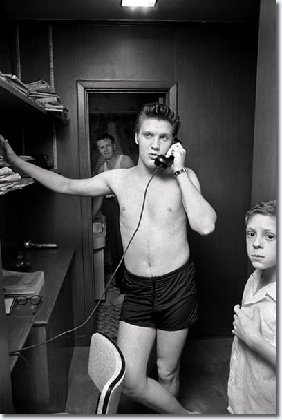 Elvis on the phone at his parents’ house with his young cousin, Billy Smith, who would later become part of the “Memphis Mafia,” Elvis’s entourage. In the background is Elvis’s father, Vernon Presley, shaving.